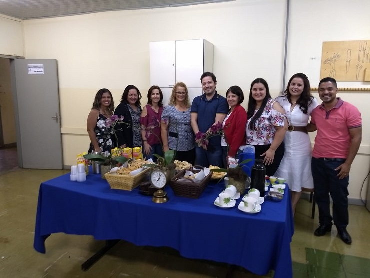 Na sala de defesa do Programa, de pé, atrás de uma mesa decorada com flores, com forro azul e com um rico café da manhã, encontram-se, respectivamente da esquerda para a direita, Tattiana (egressa), Luciene (professora), Marta (professora), Elismar (egressa), Ulysses (egresso), Flomar (professora), Caroline (egressa), Lílian (egressa), Thiago (egresso). Todos estão vestidos informalmente. 